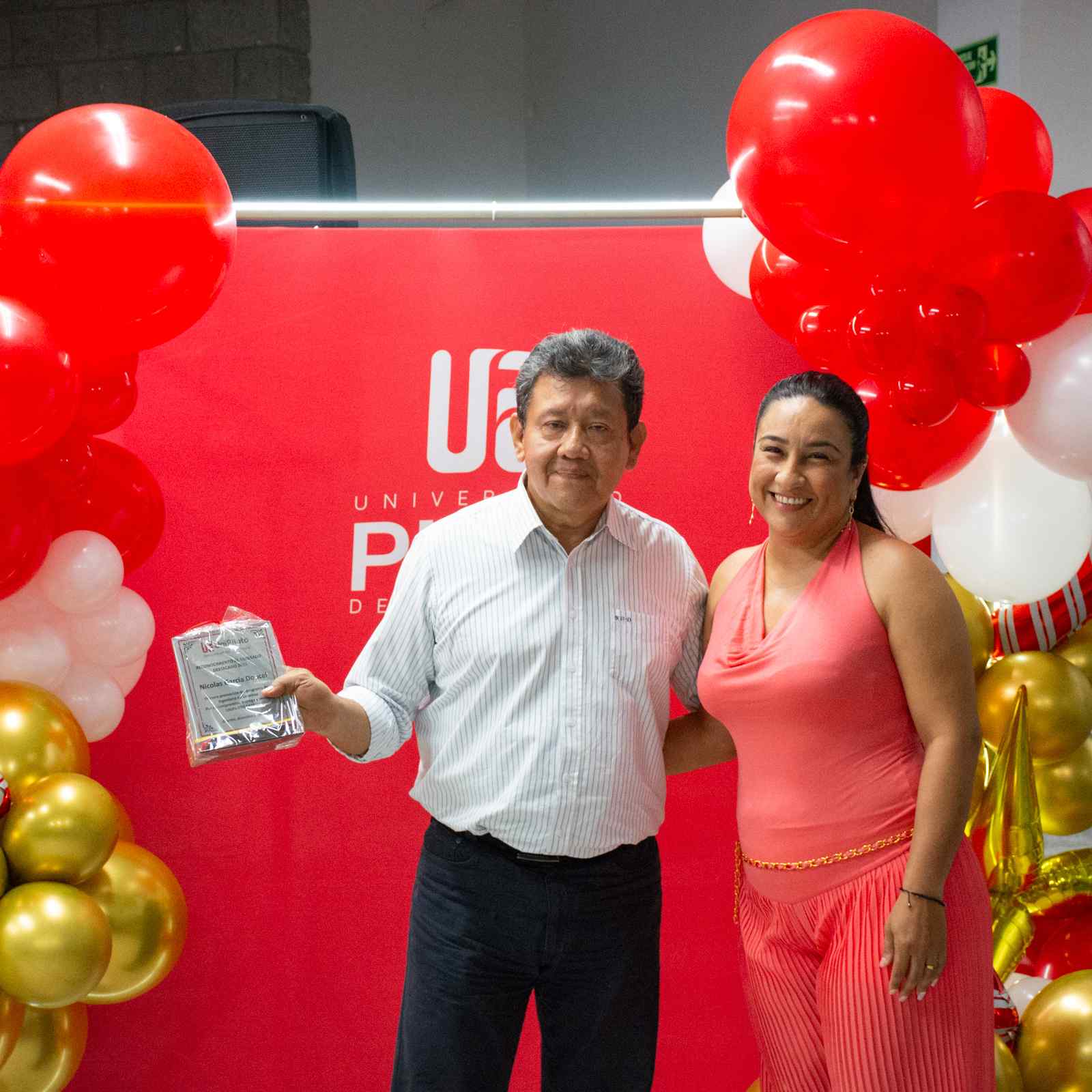 Una foto que muestra a Nicolás García Doncel (izquierda) y a Nataly Beltrán Barco (derecha). Posando sonrientes exhibiendo el reconocimiento que recibió el egresado Nicolás. Ambos están vestidos de gala, frente a un fondo rojo con el logo de la unipiloto.
