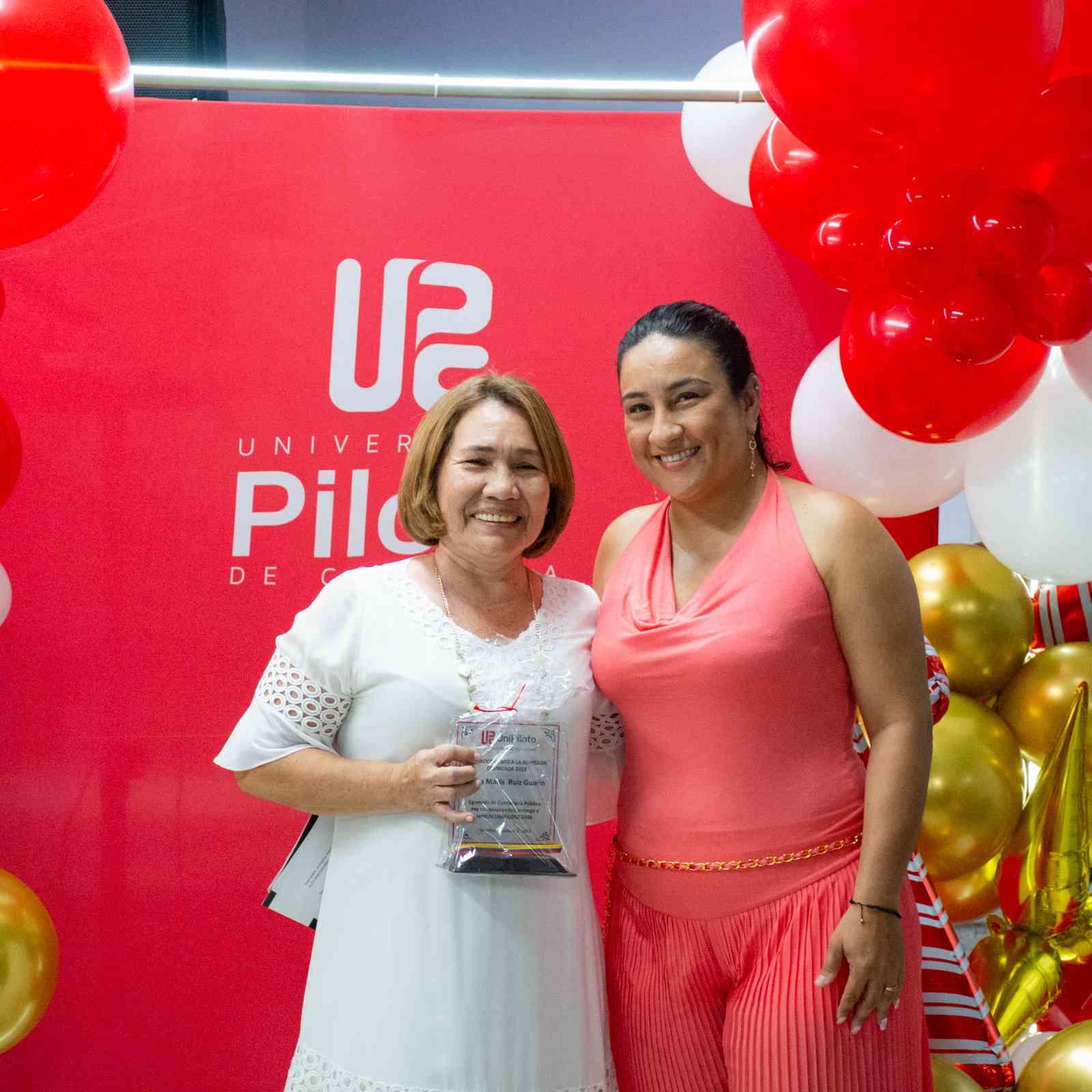 Una foto que muestra a Diana María Ruiz(izquierda) y a Nataly Beltrán Barco (derecha). Posando sonrientes exhibiendo el reconocimiento que recibió el egresado Nicolás. Ambas están vestidas de gala, frente a un fondo rojo con el logo de la unipiloto.