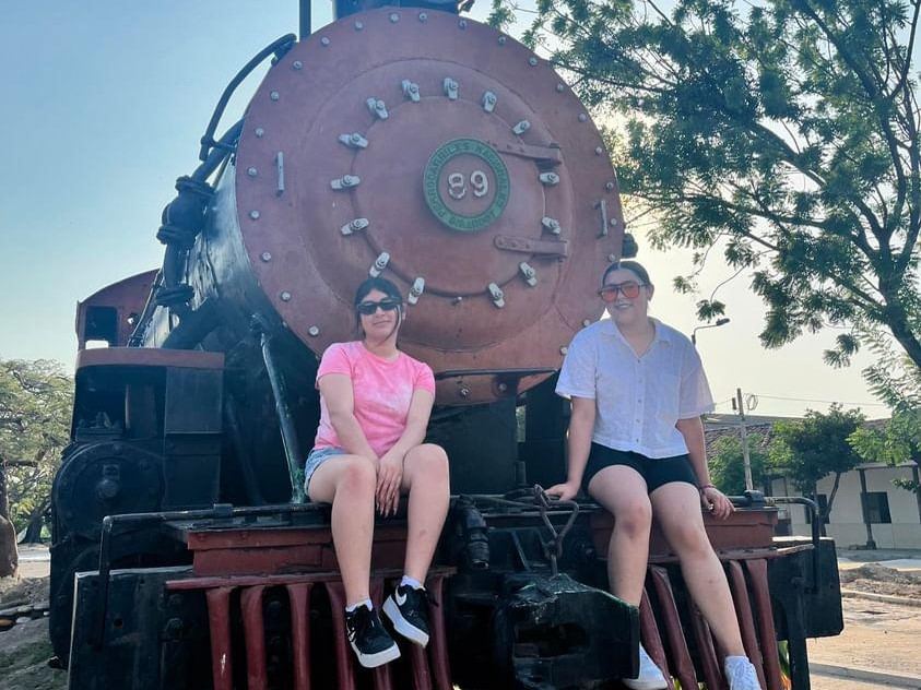 Una fotografía que muestra a las estudiantes de intercambio Amy Espino Mendoza y Karen L. Álvarez Gómez, de la Universidad Autónoma de Zacatecas. Están sentadas en el monumento al tren de Girardot, ubicado cerca a la Casa de la Cultura. Ambas se ven alegres, con un árbol en el fondo y el día se nota claro.