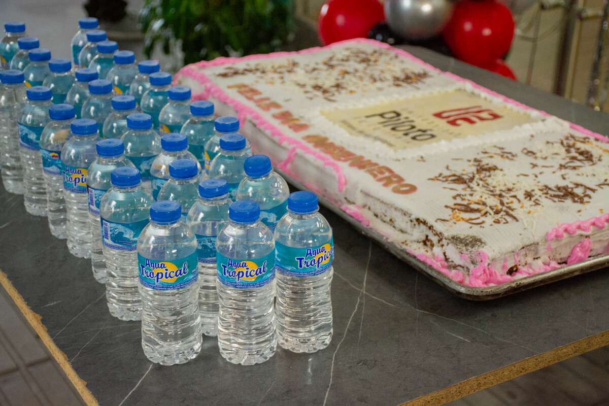 Una foto donde se observa una mesa. Sobre la mesa hay una torta grande con el logo de la Universidad, alrededor de la torta hay unas botellas con agua marga Agua Tropical.