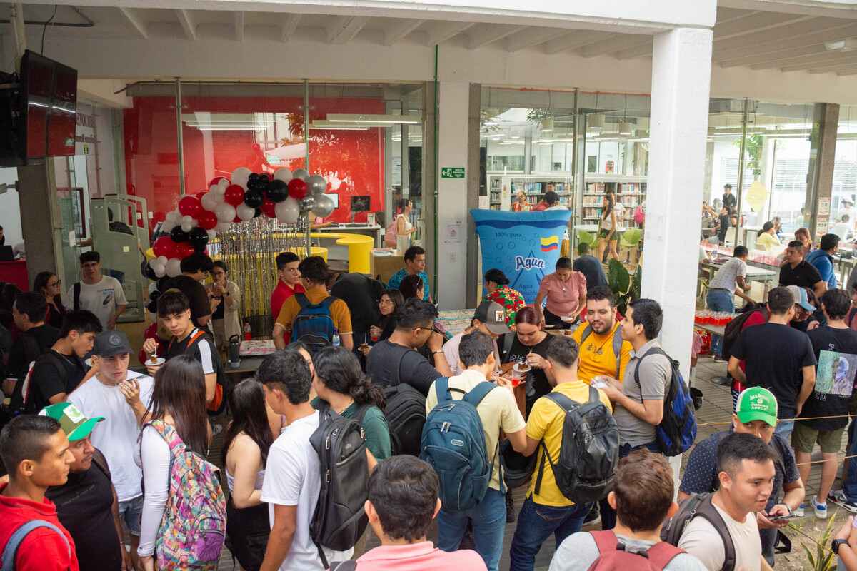 Una fotografía donde se ve el pasillo de la biblioteca de la universidad, por el pasillo están caminando muchas personas, compartiendo un momento de charla. Algunos están disfrutando mientras comen torta. De fondo se observa la biblioteca y hemeroteca de la Universidad.